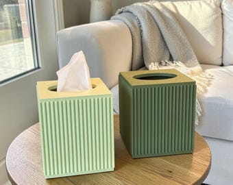 May include: Two decorative tissue box covers. One is light green, the other is olive green. Both have a square shape with vertical ribbed detailing. The light green box contains white tissues. They sit on a round wooden table.