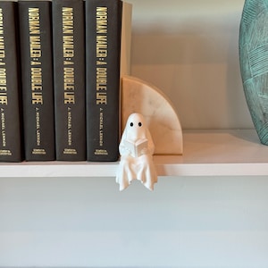 Puede incluir: Una figura de fantasma de cer&aacute;mica blanca leyendo un libro, colocada en una estanter&iacute;a blanca. El fantasma tiene dos ojos de puntos negros y est&aacute; envuelto en una s&aacute;bana fluida. Detr&aacute;s hay libros negros con letras doradas y un sujetalibros de m&aacute;rmol.