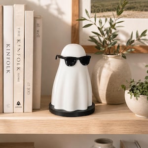 May include: A white ghost-shaped lamp wearing black sunglasses, displayed on a wooden shelf. The lamp has a black base and is positioned near a stack of books. A vase with foliage and a small potted plant are also visible.