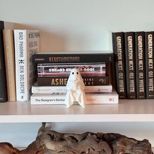 Puede incluir: Una figura de fantasma blanco de cer&aacute;mica leyendo un libro, colocada en una estanter&iacute;a con una colecci&oacute;n de libros. Los libros tienen varios t&iacute;tulos y colores, creando una exhibici&oacute;n visualmente atractiva. La estanter&iacute;a es blanca.