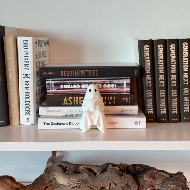 Puede incluir: Una figura de fantasma blanco de cer&aacute;mica leyendo un libro, colocada en una estanter&iacute;a con una colecci&oacute;n de libros. Los libros tienen varios t&iacute;tulos y colores, creando una exhibici&oacute;n visualmente atractiva. La estanter&iacute;a es blanca.