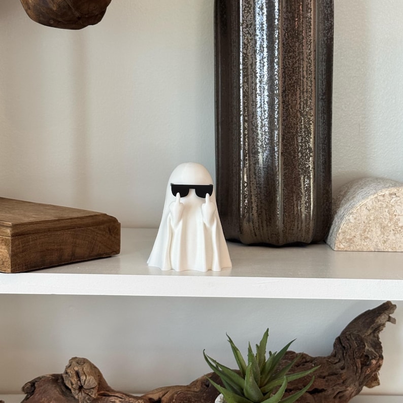 Puede incluir: Peque&ntilde;a figura de fantasma blanco con gafas de sol negras y un gesto con la mano. El fantasma est&aacute; sobre una estanter&iacute;a blanca, con un jarr&oacute;n oscuro y otros objetos decorativos en el fondo. Un adorno divertido.