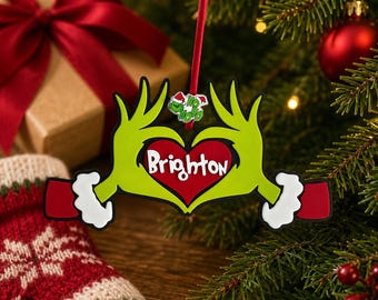 May include: A Christmas ornament featuring two green hands forming a heart shape around a red heart with the name Brighton written in white. The ornament includes a small green character icon wearing a Santa hat and the text Ho Ho Ho.
