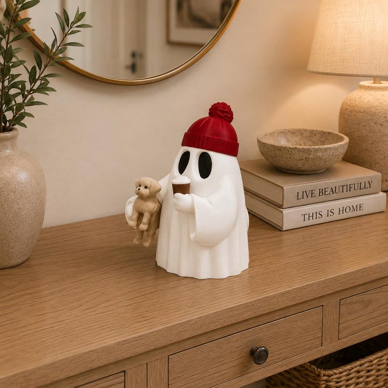 May include: A white ghost figurine wearing a red knit-style beanie with a pom-pom. The ghost has black oval eyes, holds a small brown coffee cup in one hand, and carries a small tan puppy figurine in the other hand. The figure is smooth and matte.