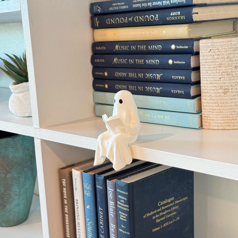 Puede incluir: Una figura de fantasma blanco leyendo un libro, posada en una estanter&iacute;a blanca. El fantasma tiene ojos negros y est&aacute; envuelto en una s&aacute;bana fluida. Libros con cubiertas azules y amarillas est&aacute;n apilados detr&aacute;s de la figura, creando un rinc&oacute;n de lectura.