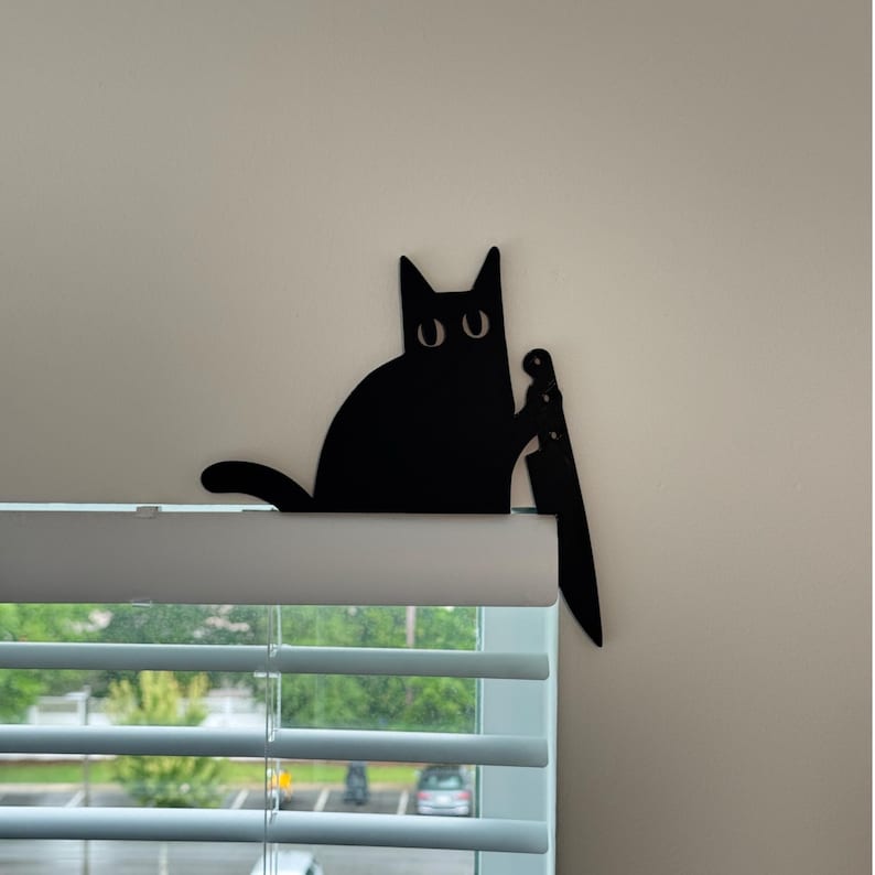 May include: A black silhouette of a cat sitting on the top edge of a window blind. The cat has large, wide eyes and is holding a large kitchen knife in one paw, with the blade pointing downward along the side of the window frame.