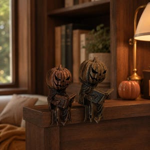 May include: Two bronze-colored pumpkin-headed figures wearing hooded cloaks, each holding an open book. The figures have carved, jagged facial features resembling jack-o'-lanterns and are positioned in a seated pose with legs dangling downward.