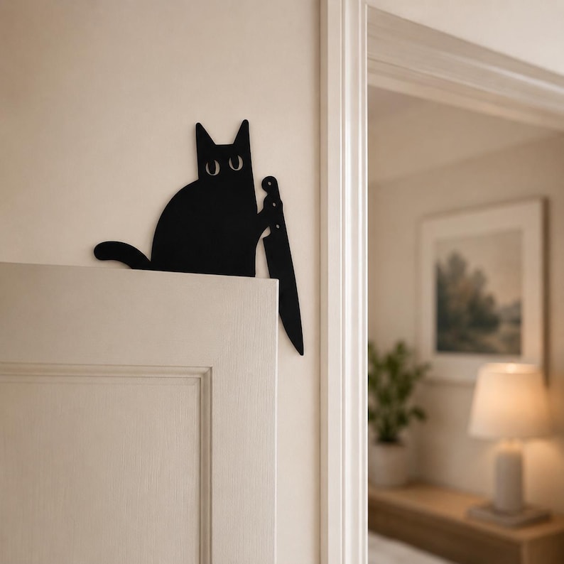 May include: A black metal wall art piece shaped like a cat silhouette holding a large kitchen knife. The cat has two simple white oval eyes and is positioned to appear as if it is peeking over the top edge of a door frame or wall corner.