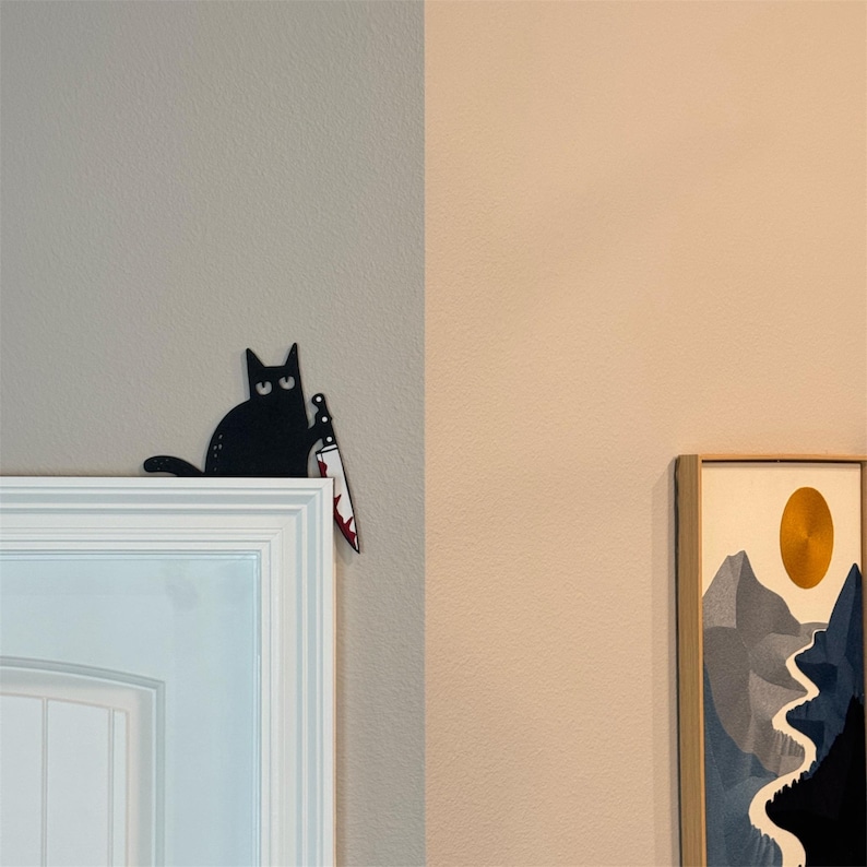 May include: A black cat wall decoration featuring a cartoon-style feline with wide eyes holding a silver knife with red blood-like stains on the blade. The cat is positioned to appear as if it is peeking over the top corner of a white door frame.