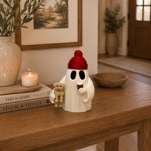 May include: A white ghost figurine wearing a red knit-style beanie, holding a small brown coffee cup in one hand and a miniature tan puppy in the other. The ghost has two large, dark, oval-shaped eye openings and a smooth, rounded body shape.