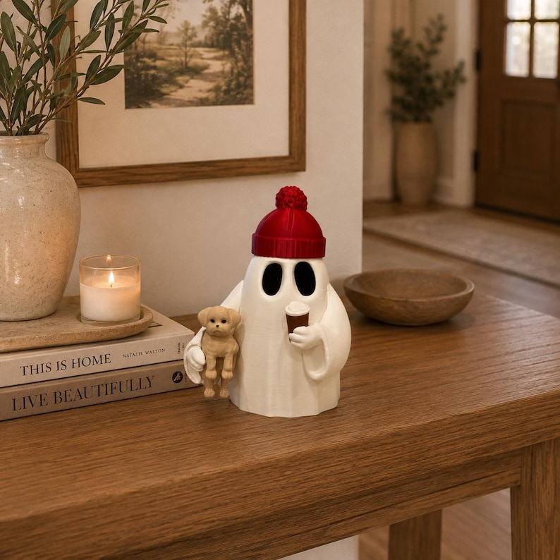 May include: A white ghost figurine wearing a red knit-style beanie, holding a small brown coffee cup in one hand and a miniature tan puppy in the other. The ghost has two large, dark, oval-shaped eye openings and a smooth, rounded body shape.