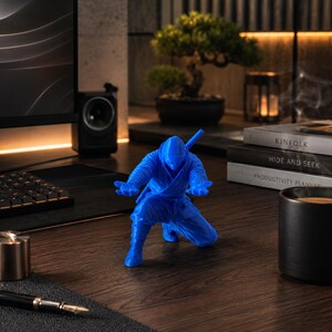 May include: A blue ninja figurine in a crouching pose with a katana on its back. The figurine is on a wooden desk, with a keyboard, monitor, and books in the background. A black coffee mug and a pen are also on the desk.