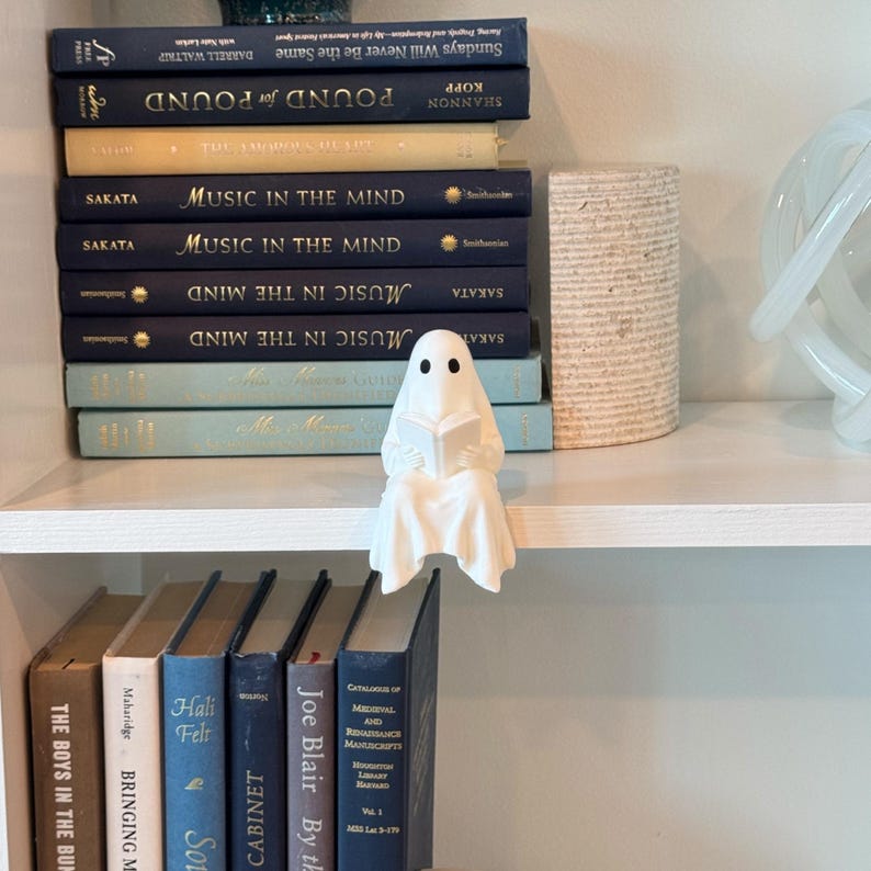 Puede incluir: Una figura de fantasma blanco leyendo un libro, sentada en una estanter&iacute;a blanca. El fantasma tiene dos ojos negros y est&aacute; envuelto en una s&aacute;bana blanca. Detr&aacute;s del fantasma hay varios libros con cubiertas azules y beige. A la derecha hay un objeto cil&iacute;ndrico decorativo.