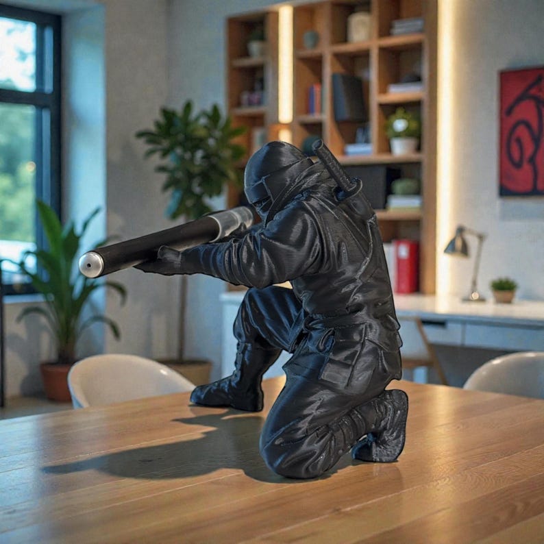 May include: A black ninja figurine in a kneeling position, aiming a long, cylindrical weapon. The figure is detailed with a helmet, gloves, and tactical clothing. A katana is strapped to the back. The figurine is on a wooden table.