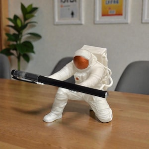 Może przedstawiać: Biała figurka astronauty trzymająca czarny długopis. Astronauta klęczy, z pomarańczową osłoną wizjera. Długopis jest czarny ze srebrną końcówką. Figurka stoi na drewnianej powierzchni.
