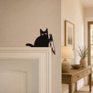 May include: A black cat wall decoration made of flat material, featuring a stylized cat with wide eyes holding a large kitchen knife with red blood-like stains on the blade. The cat is positioned to sit on the top corner of a door frame.