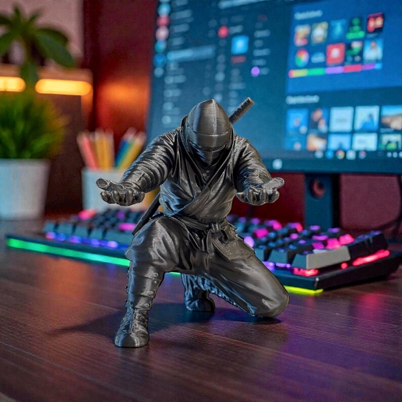 May include: A dark gray ninja figurine in a crouching pose, with arms outstretched. The ninja is wearing a full suit and a mask, with a sword on its back. The figurine is on a wooden surface, with a colorful keyboard and computer monitor in the background.