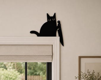 May include: A black metal wall decoration shaped like a cat holding a large kitchen knife. The cat silhouette features two circular cutouts for eyes and is positioned to appear as if it is peeking over the top corner of a window frame.