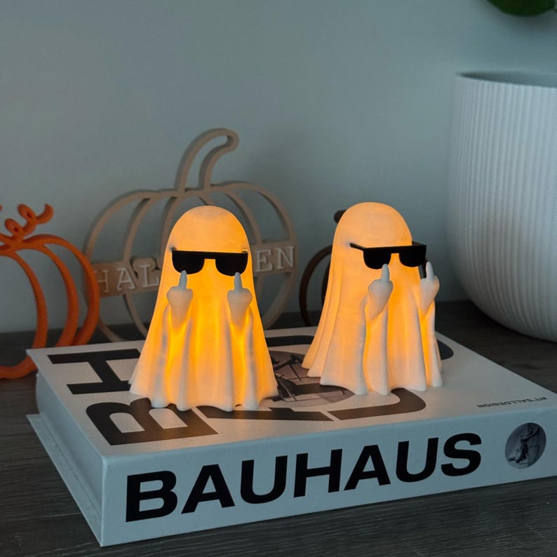 Puede incluir: Dos figuras de fantasmas luminosos con gafas de sol negras mostrando el dedo coraz&oacute;n. Est&aacute;n sobre un libro con la palabra "BAUHAUS" impresa. En el fondo, una calabaza de madera y un letrero de "HALLOWEEN".