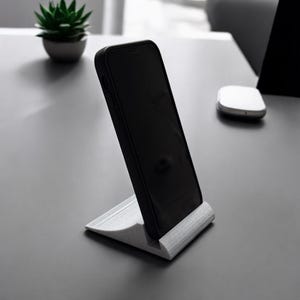 May include: A white phone stand with a black smartphone resting on it. The stand is made of a textured material and has a curved design.