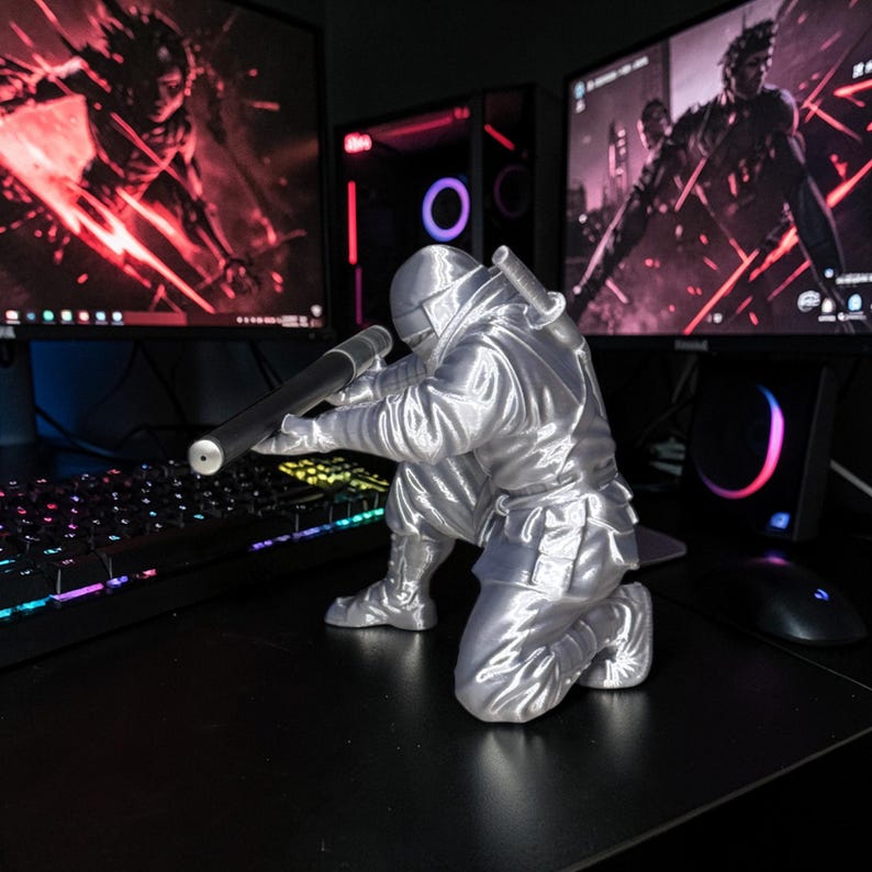 May include: A silver-colored figurine of a kneeling ninja, aiming a rifle. The figure is detailed with a helmet, uniform, and a sword on its back. The figurine is on a black desk, with computer monitors in the background.
