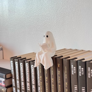 Puede incluir: Una figura de fantasma blanco leyendo un libro, posada sobre una fila de libros de tapa dura. El fantasma tiene agujeros negros para los ojos y est&aacute; envuelto en una s&aacute;bana fluida. Los libros tienen cubiertas marrones con texto blanco.