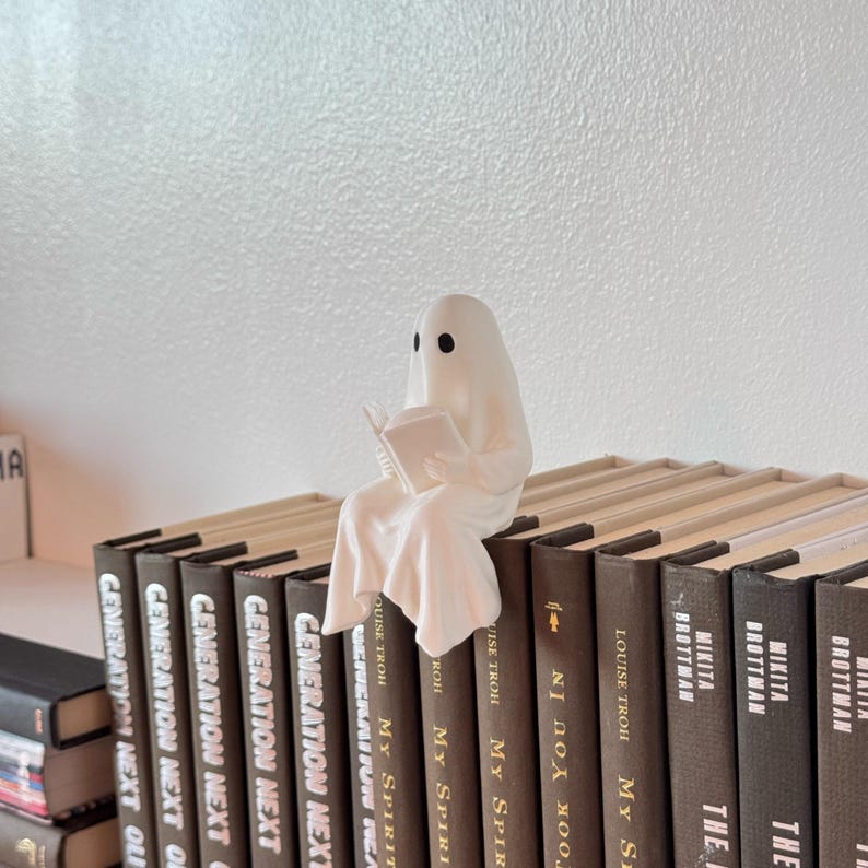 Puede incluir: Una figura de fantasma blanco leyendo un libro, posada sobre una fila de libros de tapa dura. El fantasma tiene agujeros negros para los ojos y est&aacute; envuelto en una s&aacute;bana fluida. Los libros tienen cubiertas marrones con texto blanco.