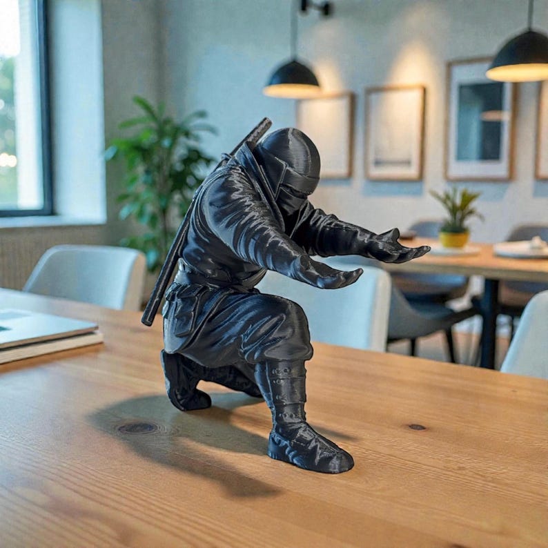 May include: A black ninja figurine in a kneeling pose with arms outstretched. The ninja is wearing a full suit with a mask and a sword on its back. The figurine is on a wooden table.