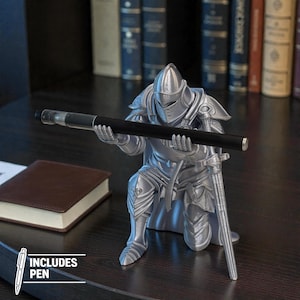 May include: A silver-colored knight figurine holding a black pen. The knight is kneeling, with a sword at its side. The image includes a brown notebook and the text "INCLUDES PEN".