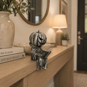 May include: A silver-toned pumpkin-headed figurine wearing a hooded robe, sitting on an edge while holding an open book. The figure features a carved jack-o'-lantern face with sharp, angular eyes and a jagged mouth, finished with a weathered texture.
