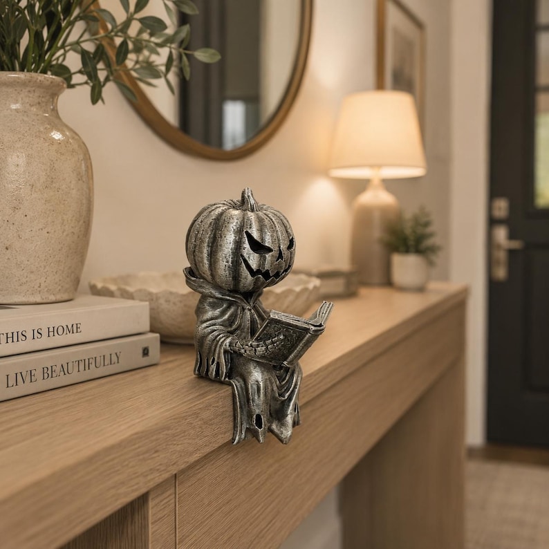May include: A silver-toned pumpkin-headed figurine wearing a hooded robe, sitting on an edge while holding an open book. The figure features a carved jack-o'-lantern face with sharp, angular eyes and a jagged mouth, finished with a weathered texture.