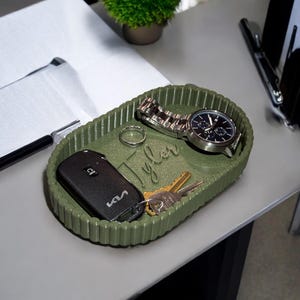May include: An oval olive green valet tray with a ribbed exterior and the name Tyler embossed in the centre. The tray holds a black Kia car key fob, a silver ring, a metal wristwatch with a link band, and two brass-coloured metal keys.