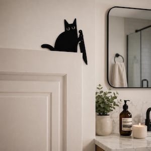 May include: A flat, black, silhouette-style wall decoration shaped like a cat with wide, white eyes. The cat is depicted in a seated position, holding a large, pointed knife in one paw, designed to be mounted on the top edge of a door frame.