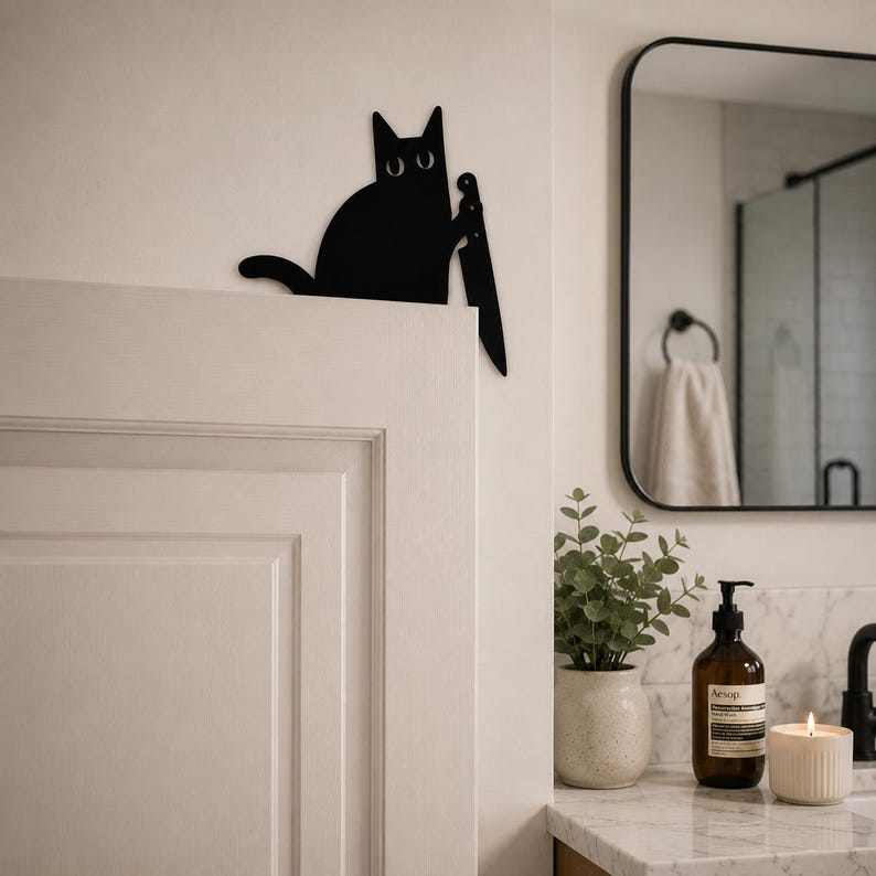 May include: A flat, black, silhouette-style wall decoration shaped like a cat with wide, white eyes. The cat is depicted in a seated position, holding a large, pointed knife in one paw, designed to be mounted on the top edge of a door frame.