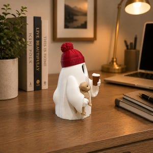 May include: A white ghost figurine wearing a red knit beanie with a pom-pom. The ghost holds a small brown puppy in one arm and a brown coffee cup in the other hand. The figurine has a smooth matte finish and is positioned upright on a flat surface.
