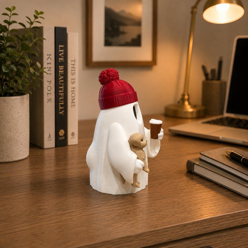 May include: A white ghost figurine wearing a red knit beanie with a pom-pom. The ghost holds a small brown puppy in one arm and a brown coffee cup in the other hand. The figurine has a smooth matte finish and is positioned upright on a flat surface.