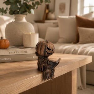 May include: A brown, carved-style figurine of a pumpkin-headed character wearing a hooded robe and sitting on the edge of a surface while holding an open book. The figure has a textured, weathered appearance with a carved jack-o'-lantern face.