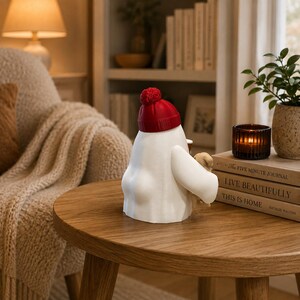May include: A white ghost figurine wearing a bright red knit-style beanie with a pom-pom on top. The ghost is holding a small brown mug and a tiny tan teddy bear. The figurine has a smooth, rounded body and is positioned on a wooden surface.