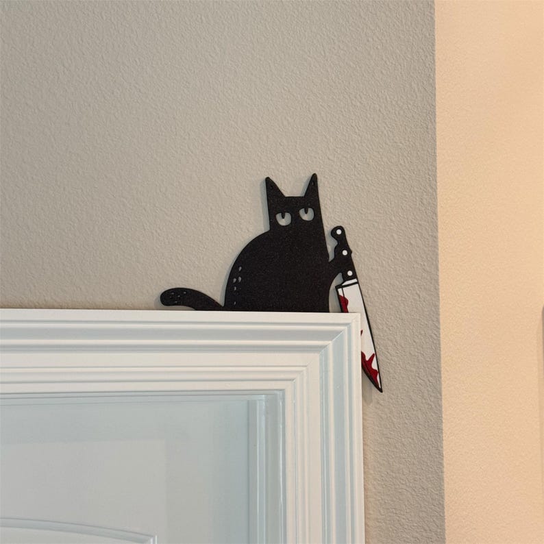 May include: A black cat wall decoration featuring a flat, silhouette-style design with wide eyes. The cat is holding a silver-colored knife with red blood-like splatters on the blade, positioned to hang on the corner of a door frame.