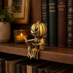 May include: A gold-toned figurine of a pumpkin-headed creature wearing a hooded robe and sitting on the edge of a shelf. The figure is holding an open book in its hands. The pumpkin head features carved, jagged eyes and a wide, toothy grin.