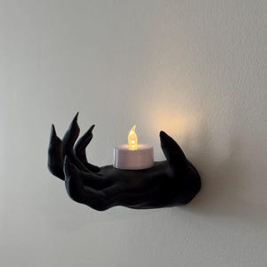 May include: A black hand-shaped wall sconce holding a small, white, battery-operated candle with a flickering yellow light. The sconce is mounted on a white wall, creating a spooky, gothic aesthetic.