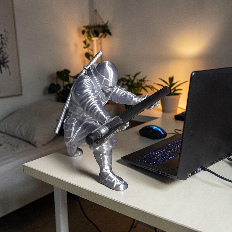 May include: A silver-colored figurine of a ninja in a crouching position, holding a black object, with a laptop and a mouse on a white desk. The ninja has a sword on its back. The scene is lit by warm light.