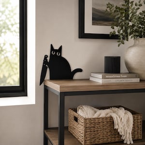 May include: A flat, black metal silhouette of a cat holding a large knife in its paw. The cat has a rounded body, pointed ears, and wide, white-outlined eyes. The design is a single-piece cutout intended to be placed on a flat surface edge.