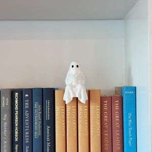 Puede incluir: Una figura de fantasma blanco leyendo un libro, posada sobre una fila de libros. Los libros son de varios colores, incluyendo azul, naranja y rojo. La figura tiene dos ojos negros y est&aacute; cubierta con una s&aacute;bana blanca.