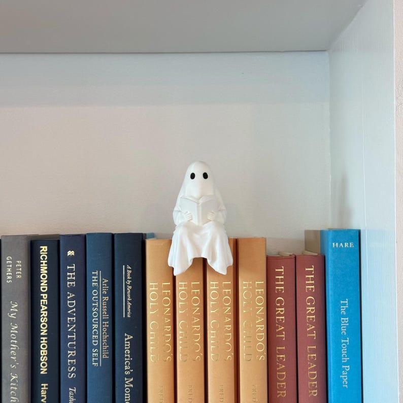 Puede incluir: Una figura de fantasma blanco leyendo un libro, posada sobre una fila de libros. Los libros son de varios colores, incluyendo azul, naranja y rojo. La figura tiene dos ojos negros y est&aacute; cubierta con una s&aacute;bana blanca.