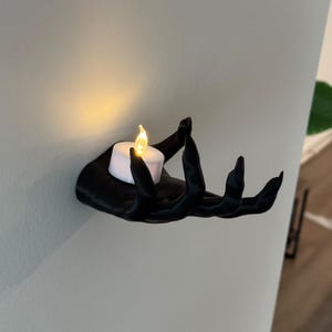 May include: A black hand-shaped wall sconce holding a white LED candle with a flickering yellow flame. The sconce is mounted on a light gray wall, creating a dramatic contrast. The candle holder is a unique decorative item.