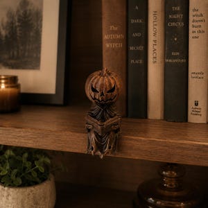 May include: A bronze-colored figurine of a pumpkin-headed creature wearing a hooded cloak and holding an open book. The figure has a carved, grinning jack-o'-lantern face with hollow eyes and mouth, and it is perched on the edge of a wooden shelf.