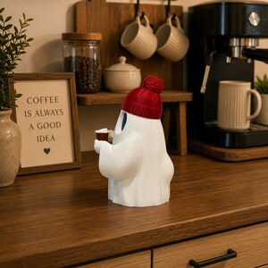 May include: A white ghost figurine wearing a red knit beanie with a pom-pom on top. The ghost has dark oval eyes and is holding a small brown coffee cup in its hands. The figurine is smooth and stands upright on a flat surface.