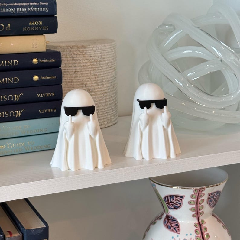 Puede incluir: Dos figuras de fantasmas blancos con gafas de sol negras y el dedo coraz&oacute;n levantado. Los fantasmas est&aacute;n sobre una estanter&iacute;a blanca, con libros y objetos decorativos en el fondo. Un art&iacute;culo de decoraci&oacute;n del hogar divertido y peculiar.