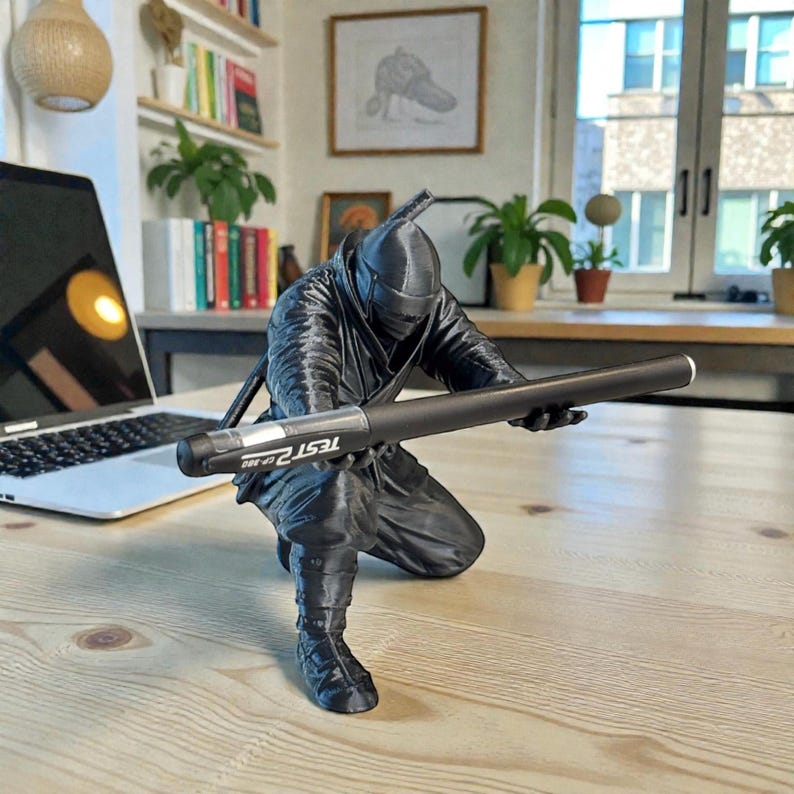 May include: A black ninja figurine in a crouching pose, holding a black pen with the text "TEST 2" on it. The ninja is wearing a full black outfit, including a mask and a sword on its back. The figurine is on a light-colored wooden surface.